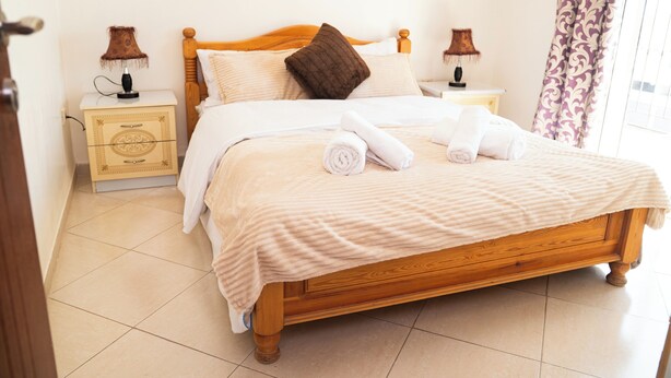 Cama bem arrumada com cabeceira de madeira, duas mesas de cabeceira com luminárias e uma janela com cortinas.