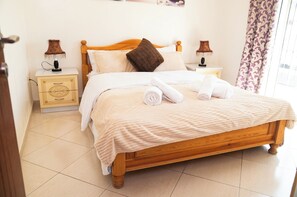 Een net opgemaakt bed met een houten hoofdbord, twee nachtkastjes met lampen en een raam met gordijnen.