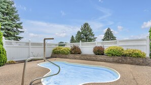 Una pequeña piscina al aire libre con una escalera metálica, rodeada por una valla blanca y arbustos diversos.