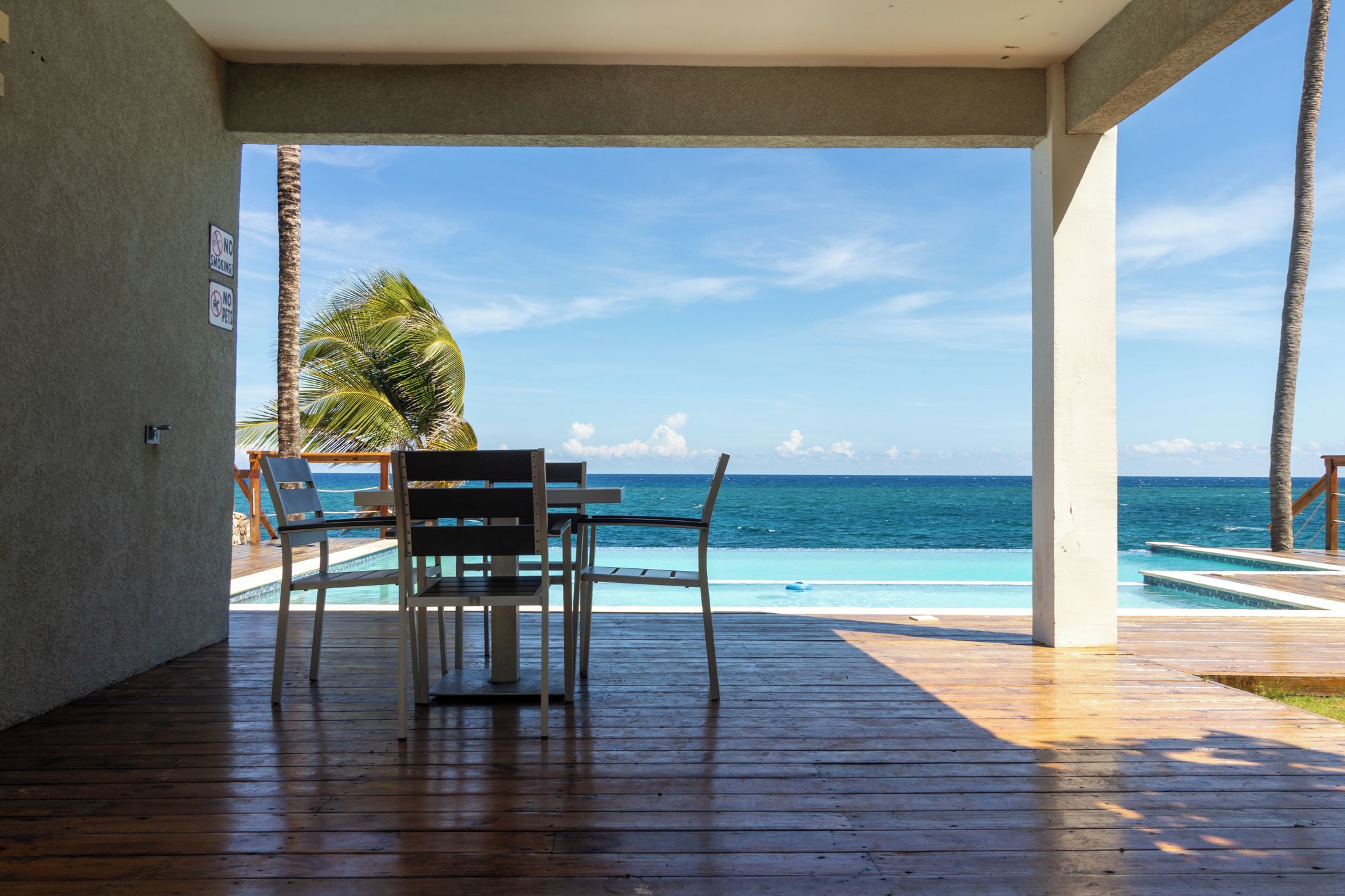 A wooden deck with a table and chairs overlooking a pool and the ocean.