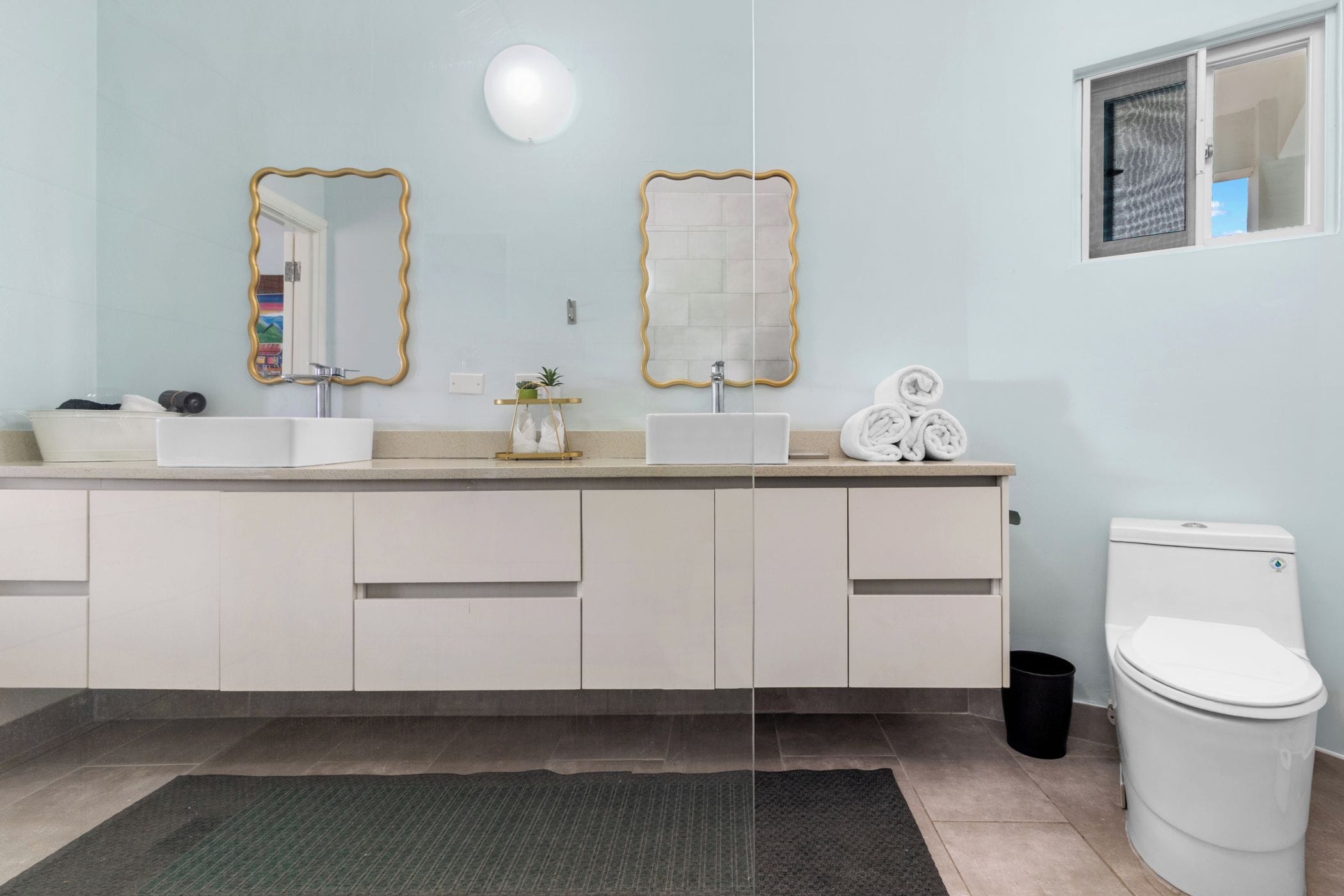 A modern bathroom with a white sink, two mirrors with gold frames, a white toilet, and a small window.