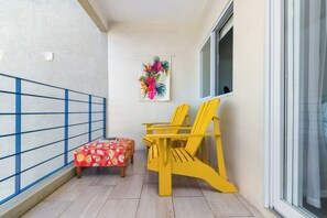 A balcony with a blue railing, a small table with a red floral tablecloth, two yellow chairs, and a wall-mounted floral tapestry. - Blue Serenity Oceanfront Penthouse (Tower Isle)