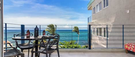 A balcony with a table and chairs, overlooking a coastal view with palm trees and the sea.