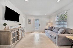 Ein modernes Wohnzimmer mit Fernseher, grauem Sofa, Holzschrank und einer Tür zu einem weiteren Raum.