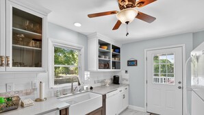 Una cocina con armarios blancos, ventilador de techo, ventana y puerta.