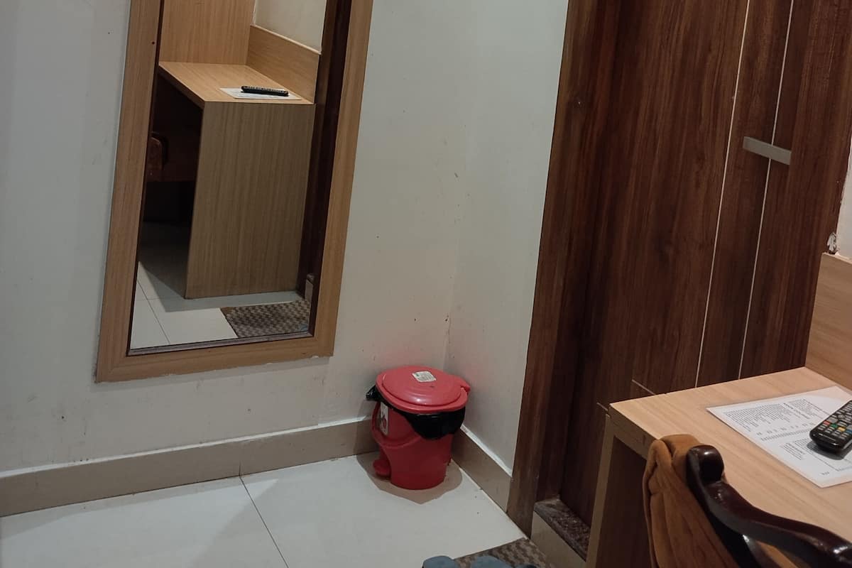 Una habitación pequeña con una puerta de madera, un espejo, un cesto de basura rojo, un escritorio con un control remoto y un par de zapatillas en el suelo.
