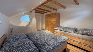 Une chambre située dans un espace de type « loft », avec un plafond en pente, des poutres en bois, un puits de lumière, un radiateur, une armoire et un lit avec une literie à motifs.