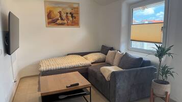 A living room with a sofa, a coffee table, a TV, a painting, a window with blinds, and a potted plant.
