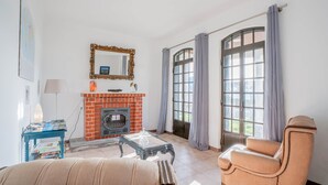 A living room with a fireplace, a sofa, a chair, a table, and a large window with curtains.