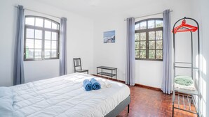 A hotel room with a bed, two windows with curtains, a chair, a small table, and a red hanger rack.