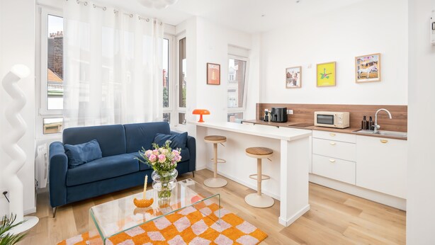 A modern living room with a blue sofa, a glass coffee table, and a kitchen area with a white countertop and bar stools. - Palm Springs - Studio Design & Lumineux in Malo Proche Plage (Dunkerque)