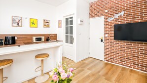 A modern kitchen with a white countertop, wooden stools, a coffee maker, a microwave, and a TV mounted on a brick wall. - Palm Springs - Studio Design & Lumineux in Malo Proche Plage (Dunkerque)