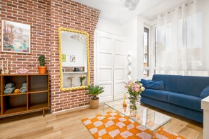 Una sala de estar con pared de ladrillo, un sofá azul, una mesa de centro de cristal y una estantería de madera con toallas.