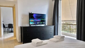 Ein Hotelzimmer mit Bett, Fernseher auf einem Nachttisch, Balkon mit Geländer und Schreibtisch mit Stühlen.
