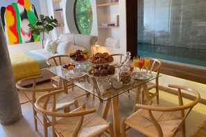 Un coin repas avec une table en verre, des chaises en osier, des aliments pour le petit-déjeuner et une vue sur un espace extérieur verdoyant.