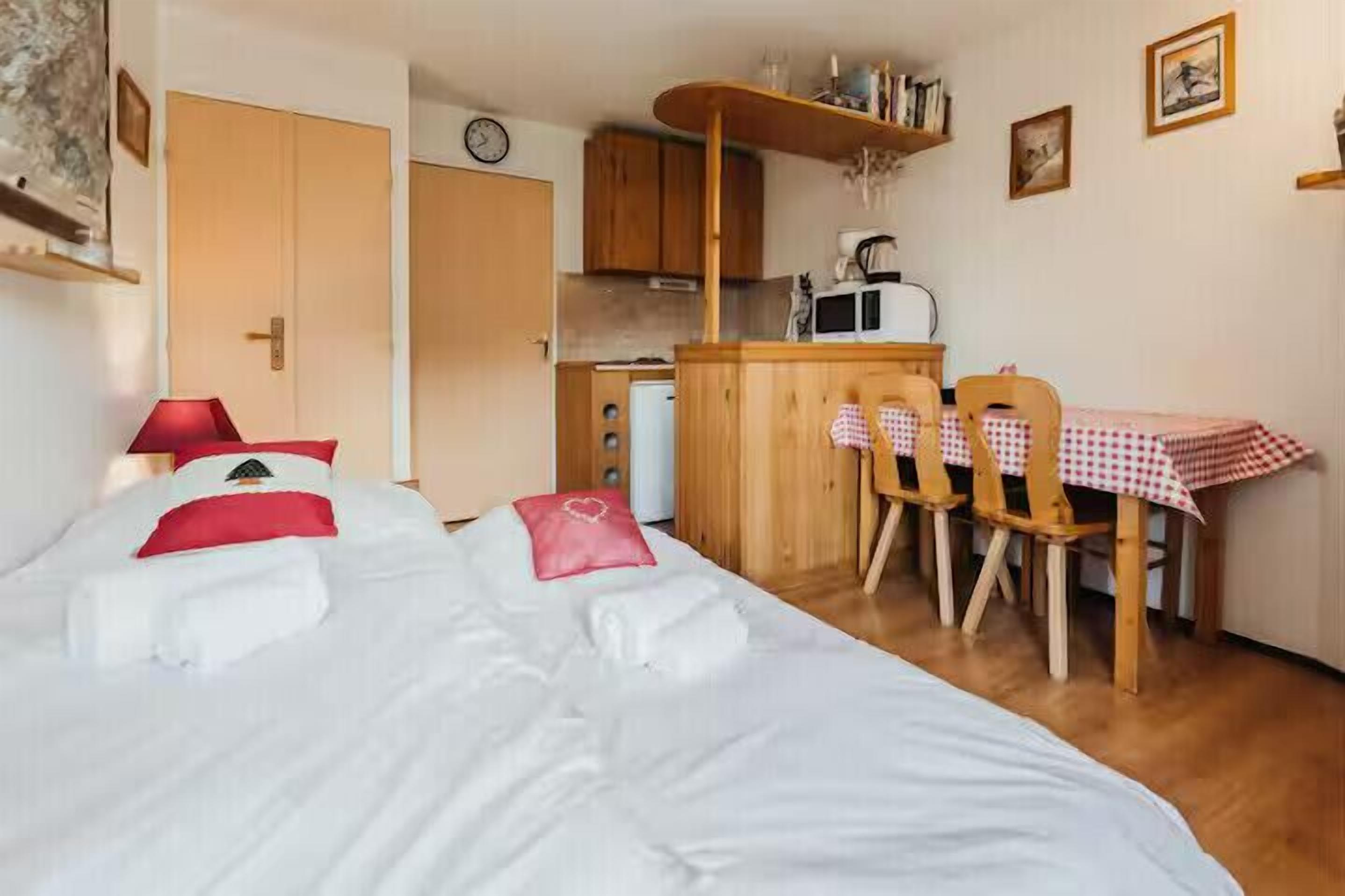A bedroom with a bed, a small kitchenette, a dining table with chairs, and a wooden cabinet.