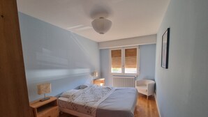 Ein Schlafzimmer mit Bett, Nachttischen, einem Sessel, einem Fenster mit Jalousien und einer Heizung.