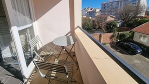 Ein Balkon mit Tisch und Stühlen, ein Fenster mit Jalousien und Blick auf eine Wohngegend mit Gebäuden und einem parkenden Auto.