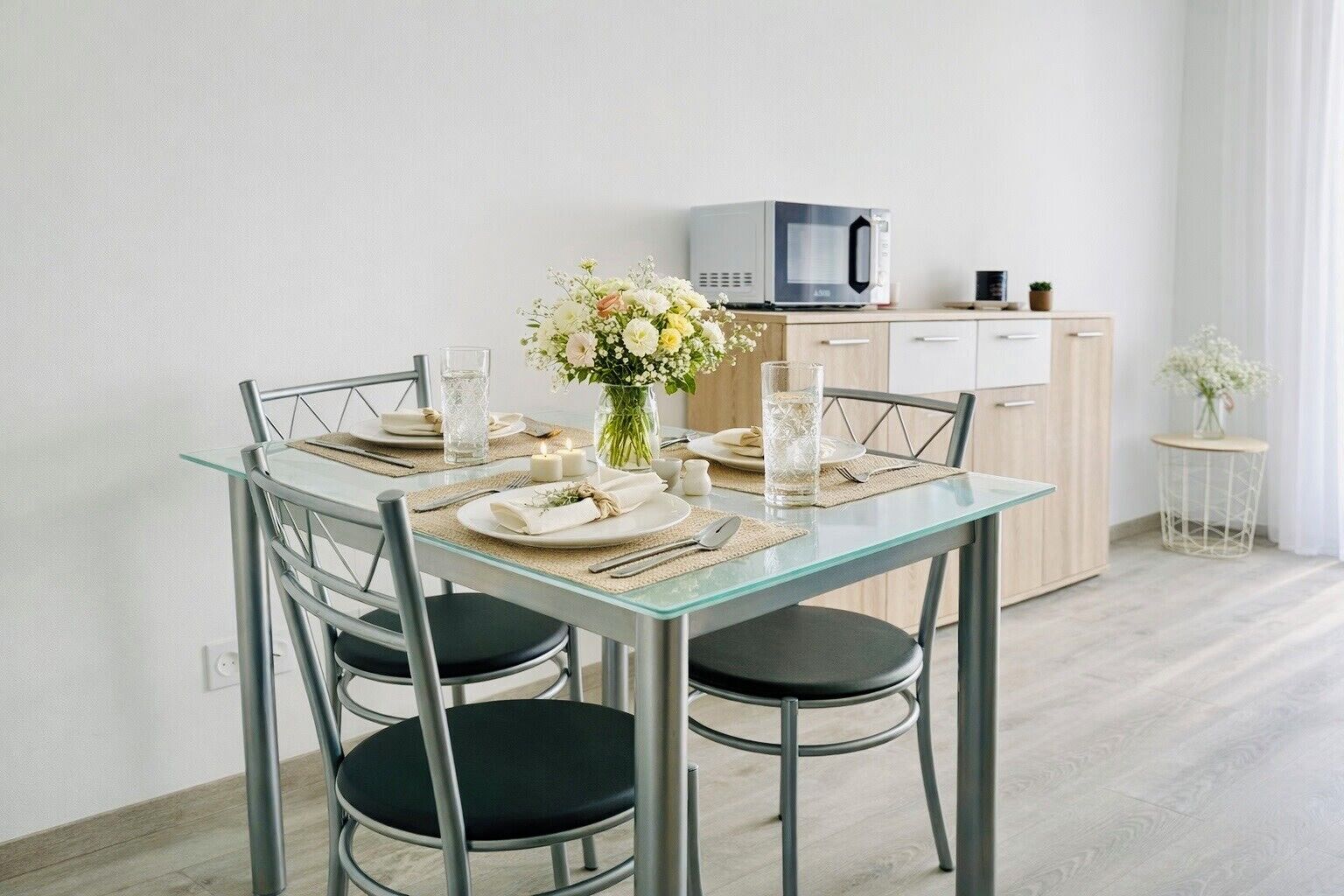 A modern dining area with a glass table, metal chairs, a vase of flowers, and a microwave in the background.
