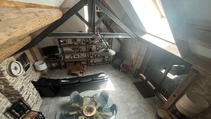 A loft-style room with exposed wooden beams, a stone wall, a fireplace, a round glass table, and a black leather sofa. - Ma Lozerienne near Banassac, A75, ideal place for a get-together. (Saint-Saturnin)
