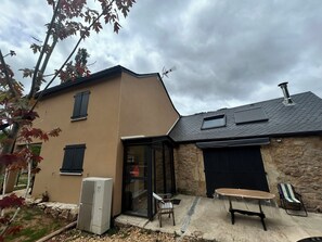 A house with a beige exterior, a stone wall, a black garage door, a small window, and a table with chairs outside. - Ma Lozerienne near Banassac, A75, ideal place for a get-together. (Saint-Saturnin)
