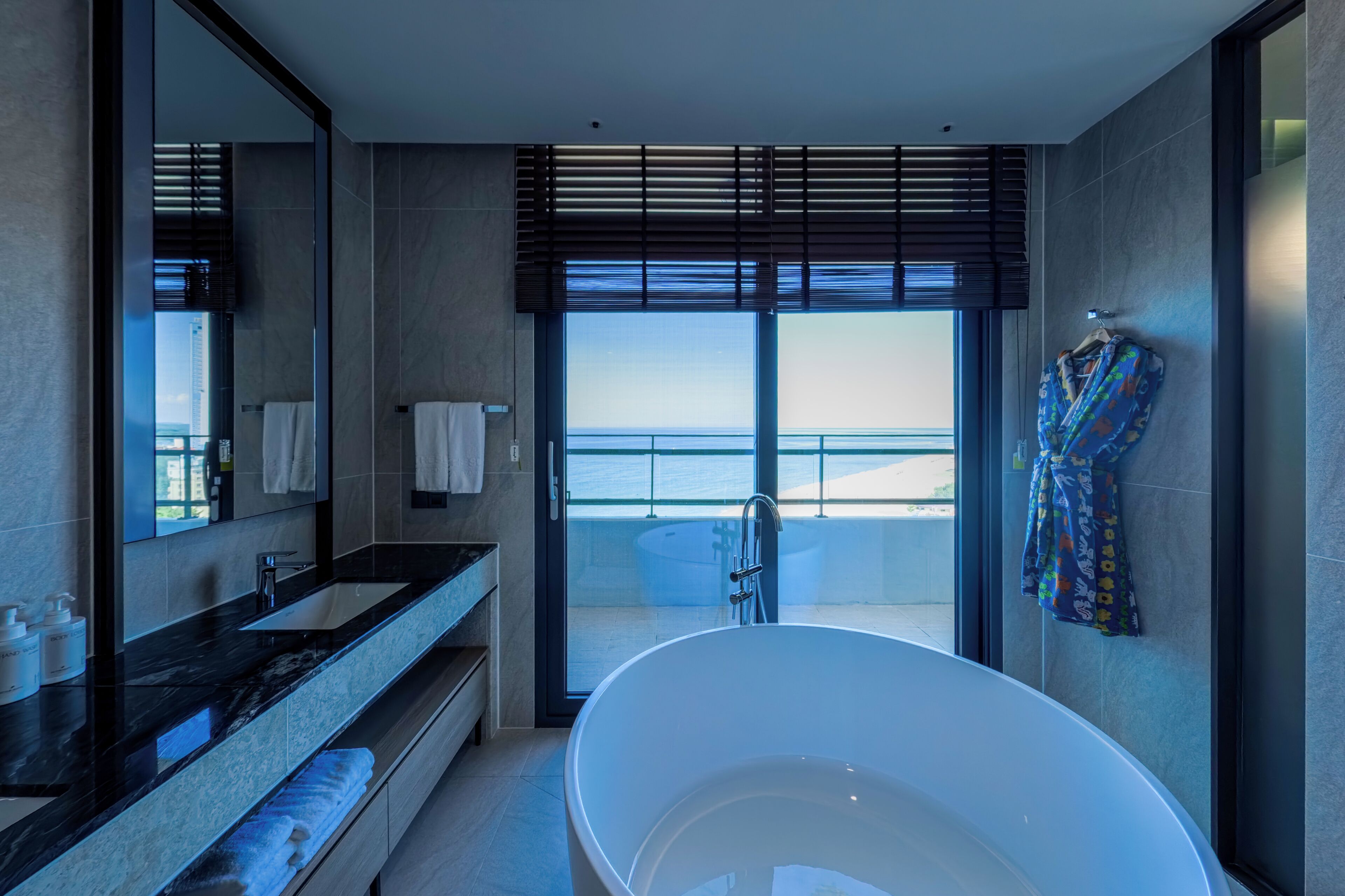 A modern bathroom with a large bathtub, a vanity with a mirror, and a view of the sea through the window.