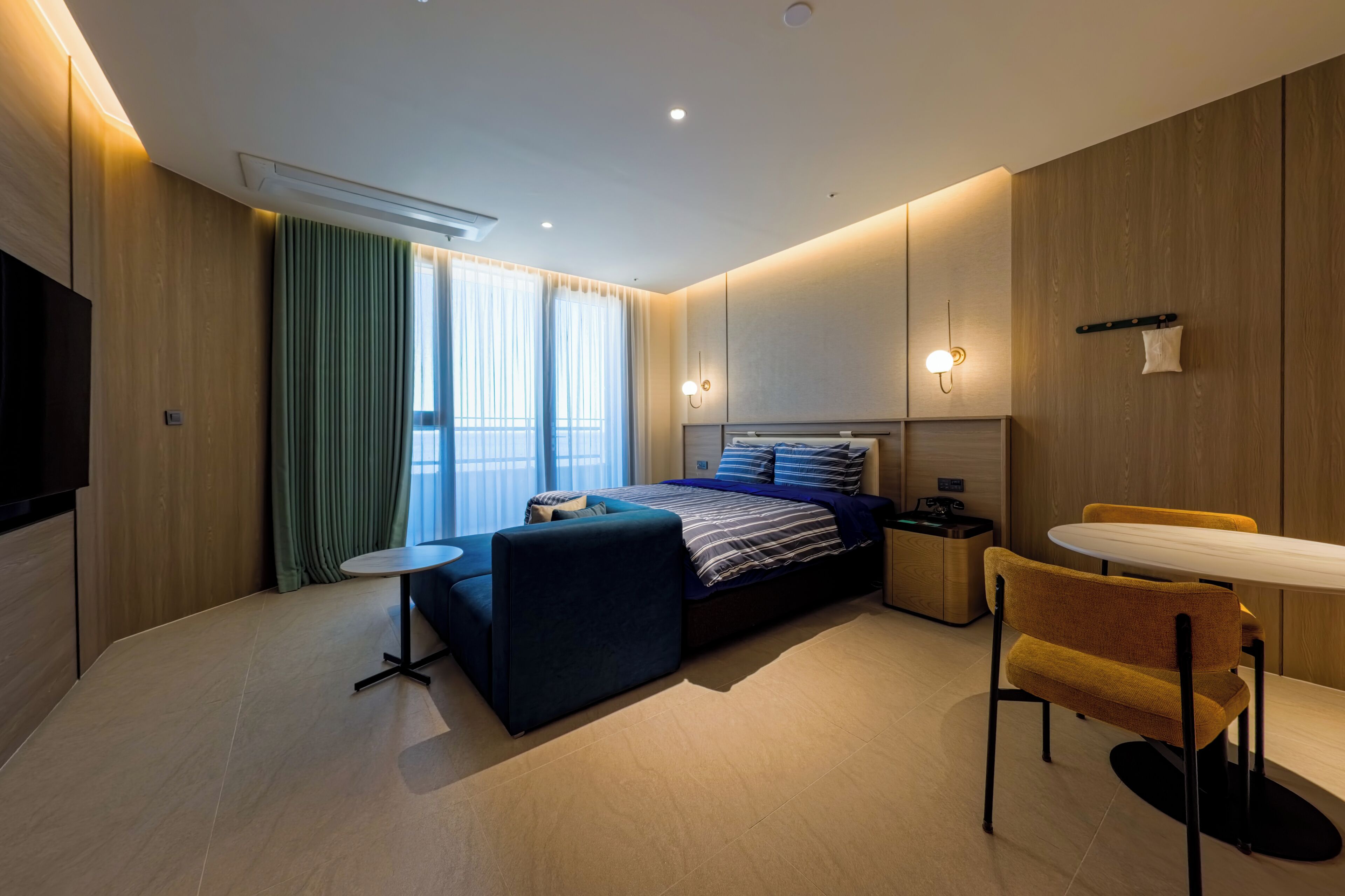 A modern hotel room with a bed, a small table, a chair, a TV, and a window with curtains.
