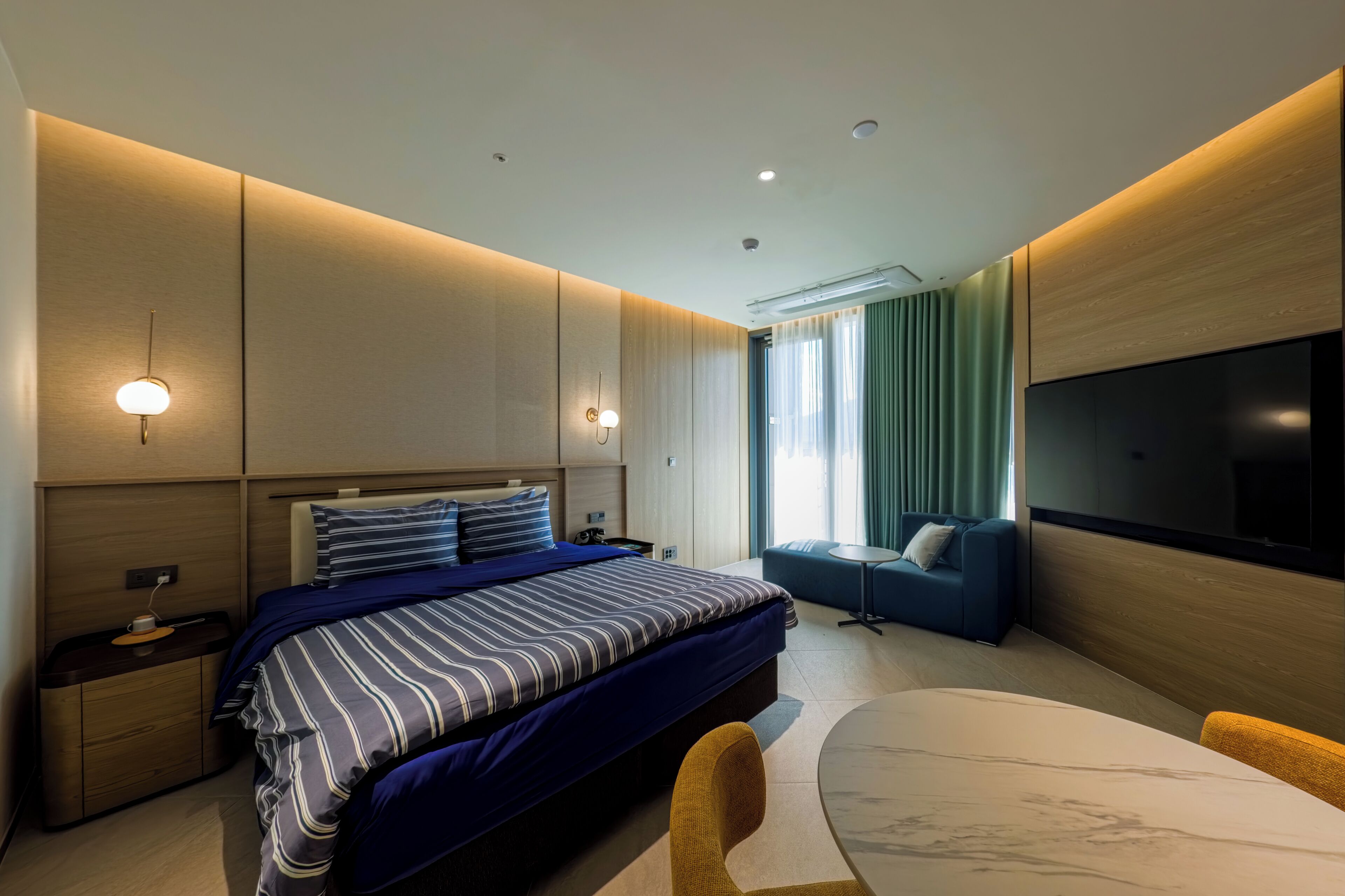 Une chambre d’hôtel moderne dotée d’un grand lit, de tables de chevet, d’une télévision, d’un coin salon et d’une grande fenêtre avec des rideaux.