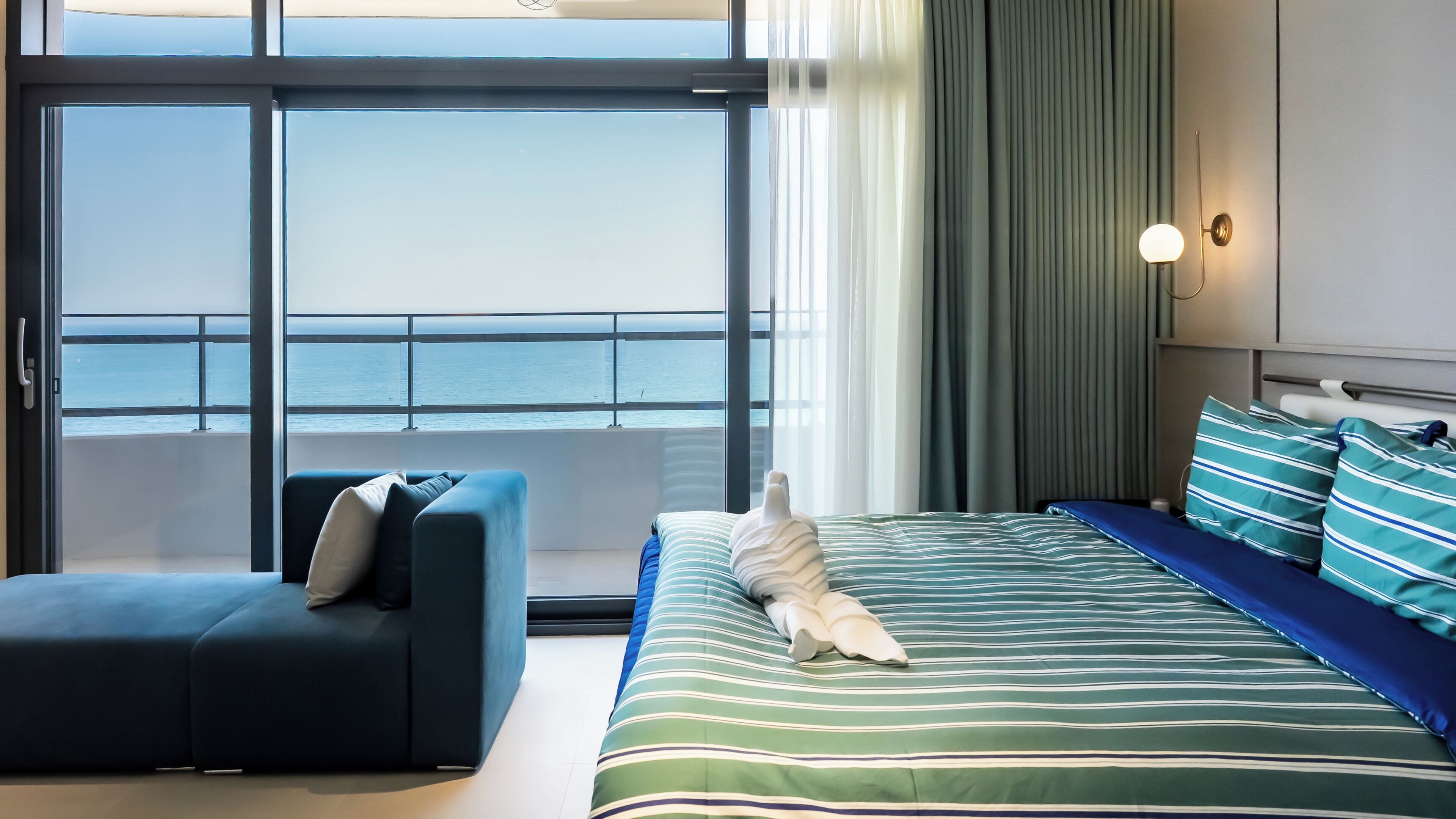 A hotel room with a large window, a bed with striped bedding, a blue sofa, and a nightstand with a lamp.