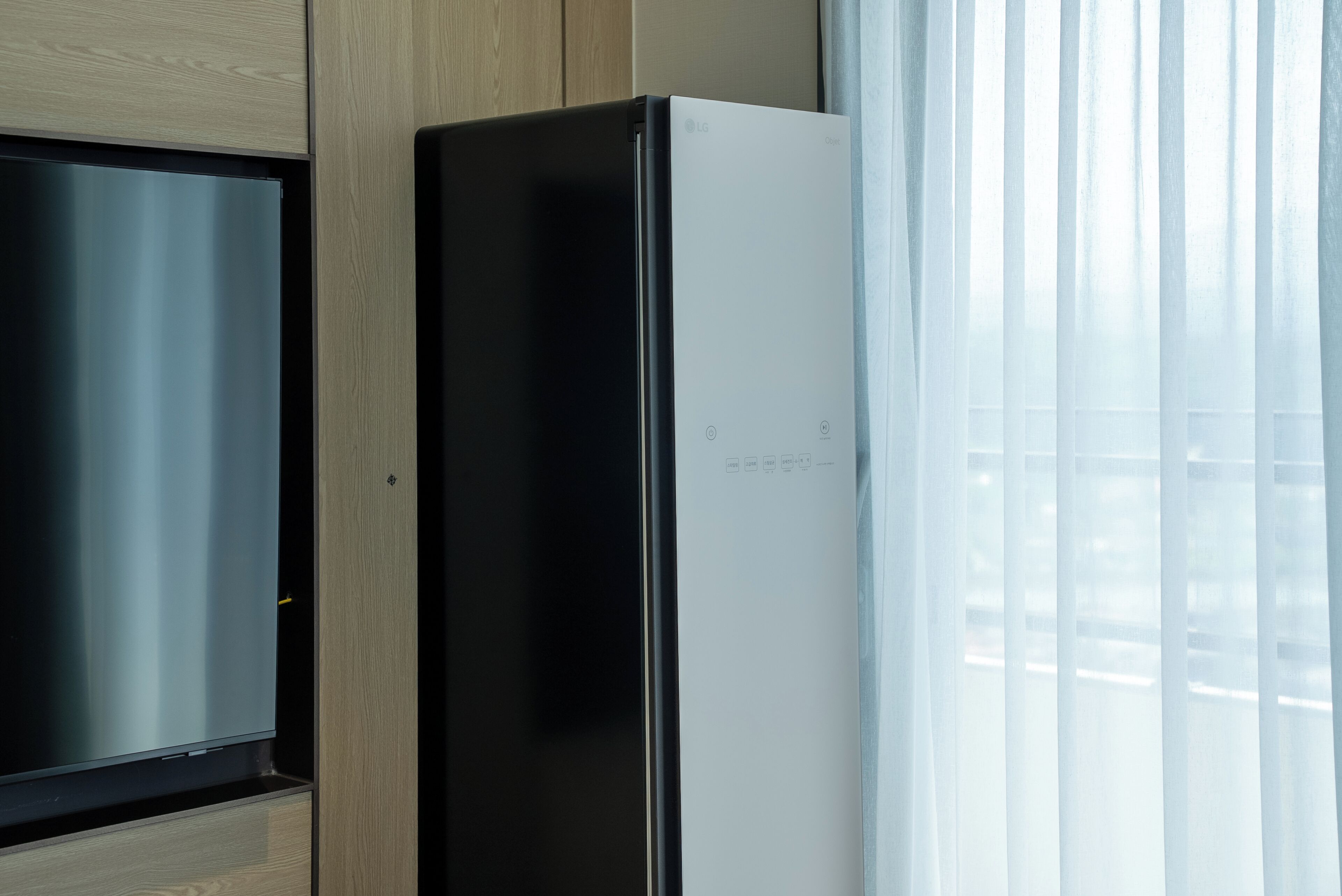 A modern kitchen with a stainless steel refrigerator, wooden cabinets, and a window with sheer curtains.