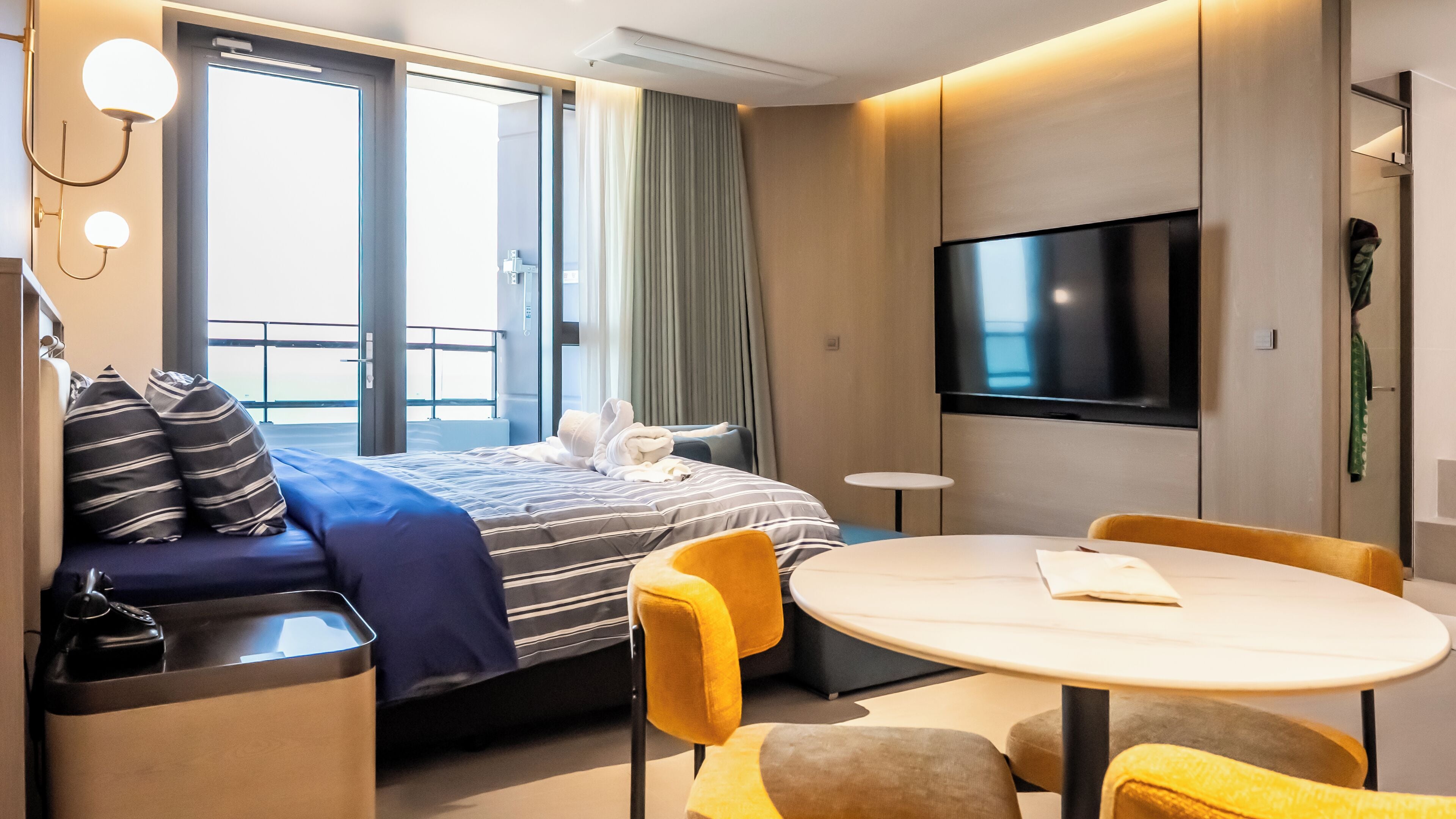 A hotel room with a bed, a TV, a dining table, and a balcony.