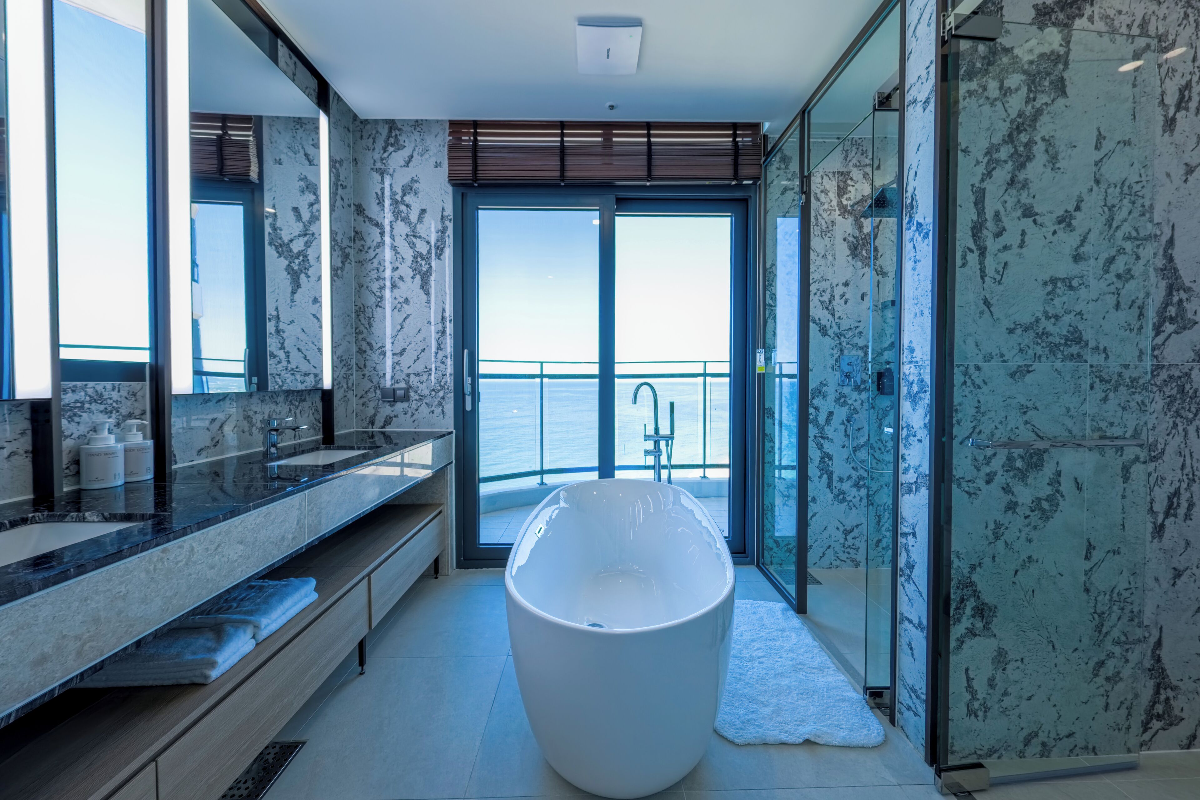 Une salle de bain moderne dotée d’une grande baignoire, d’un meuble-lavabo avec miroir et offrant une vue sur la mer grâce à une porte vitrée.