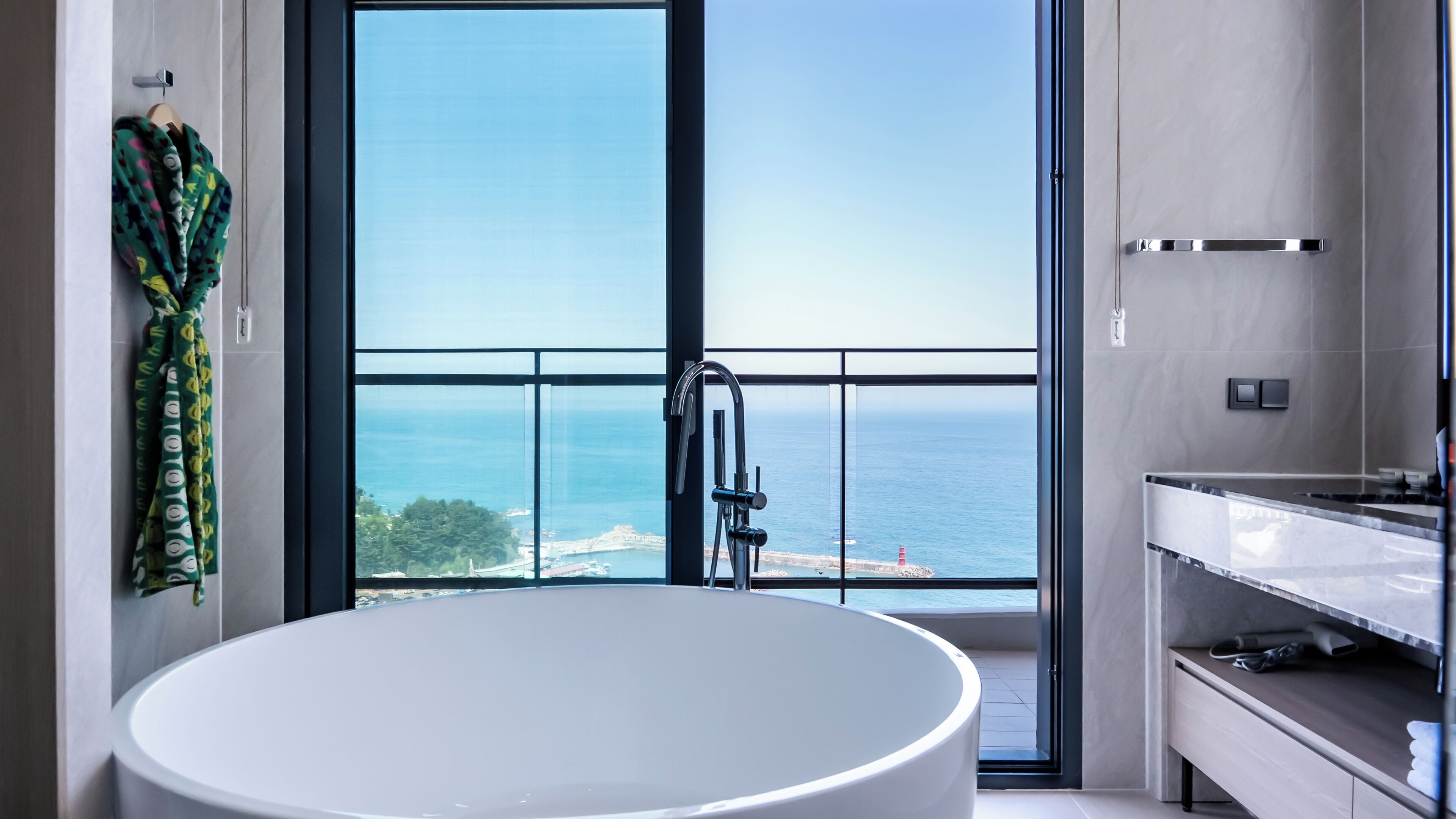 A modern bathroom with a large bathtub, a vanity with a mirror, and a view of the sea through a large window.