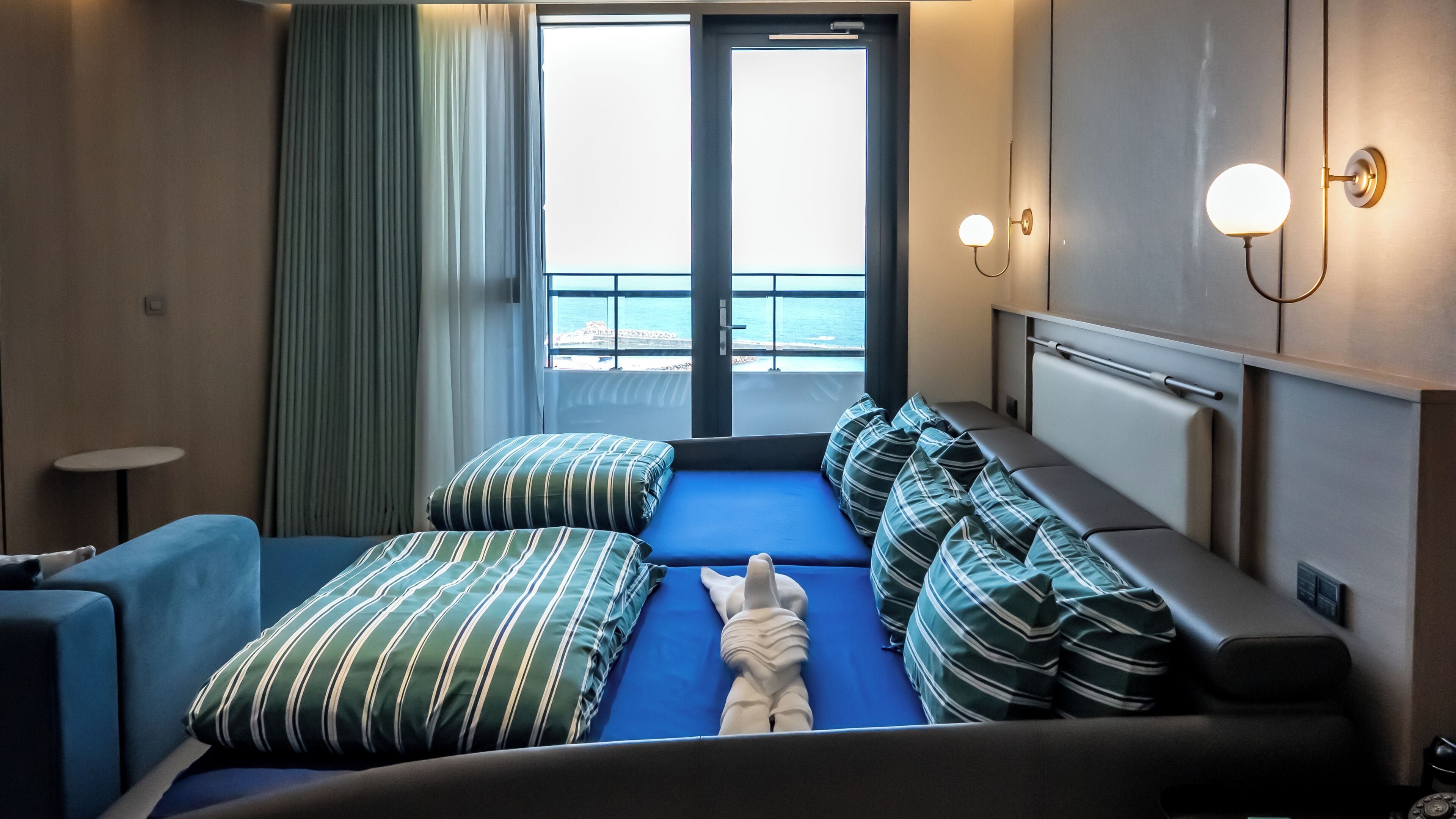 A hotel room with a bed, two chairs, a small table, a wall-mounted lamp, and a view of the sea through a large window.