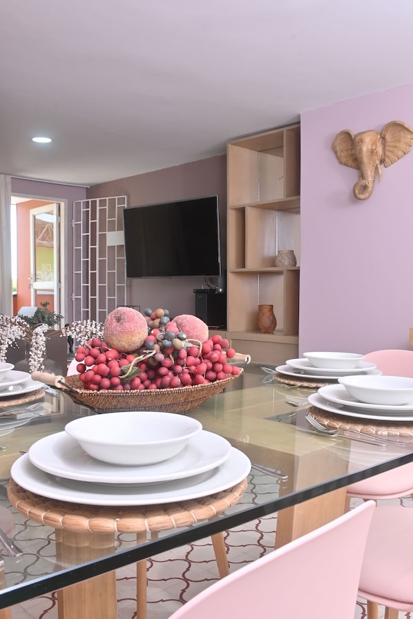 A dining area with a glass table, white plates, a fruit basket, and a wall-mounted elephant decoration.