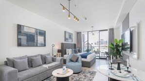 A modern hotel room with a grey sofa, a bed with a blue accent, a dining area with a table and chairs, and large windows offering a city view. - Vogue | Binghatti Galaxy JVC near Circle Mall (Dubai)