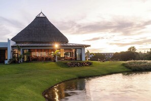 A modern building with a thatched roof, a covered outdoor seating area, and a pond. - Azuri Resort Villa: Pool, Golf & Beach Club (Rivière du Rempart)