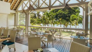 An outdoor dining area with tables and chairs, a wooden deck, and a view of the ocean and trees. - Azuri Resort Villa: Pool, Golf & Beach Club (Rivière du Rempart)