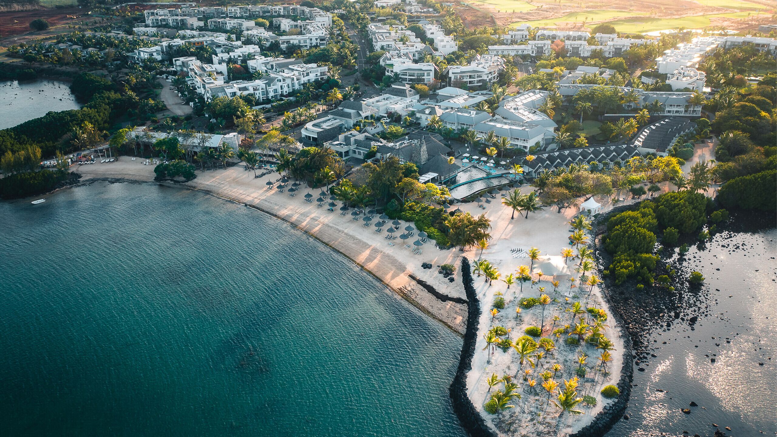 Vue aérienne d’une station balnéaire avec des villas en bord de mer, une plage de sable et une mer calme.