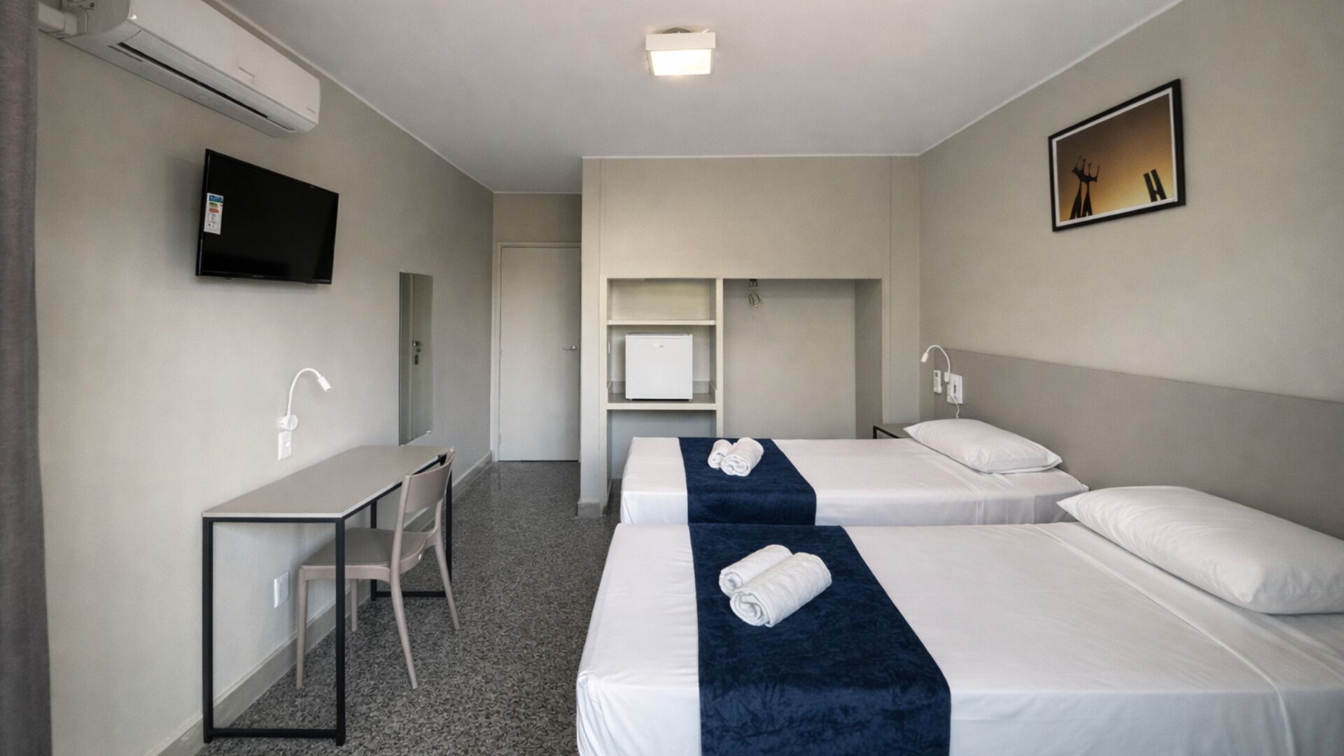 Une chambre d’hôtel avec deux lits, un bureau, une chaise, une télévision, une armoire et un tableau au mur.
