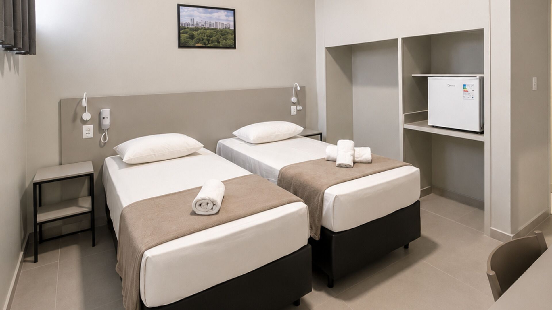 Een hotelkamer met twee bedden, een klein nachtkastje, een telefoon aan de muur, een schilderij en een ingebouwde minibar.