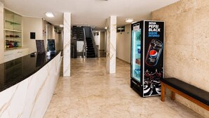 Un vestíbulo de hotel con una recepción, una máquina expendedora, una escalera y un banco.