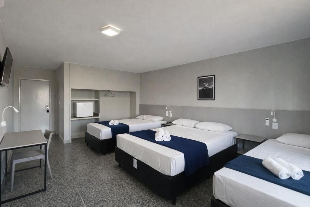 A hotel room with three beds, a desk, a chair, a TV, and a small shelf.