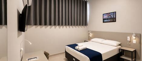 A hotel room with a bed, bedside tables, a desk, a TV, and a painting on the wall.