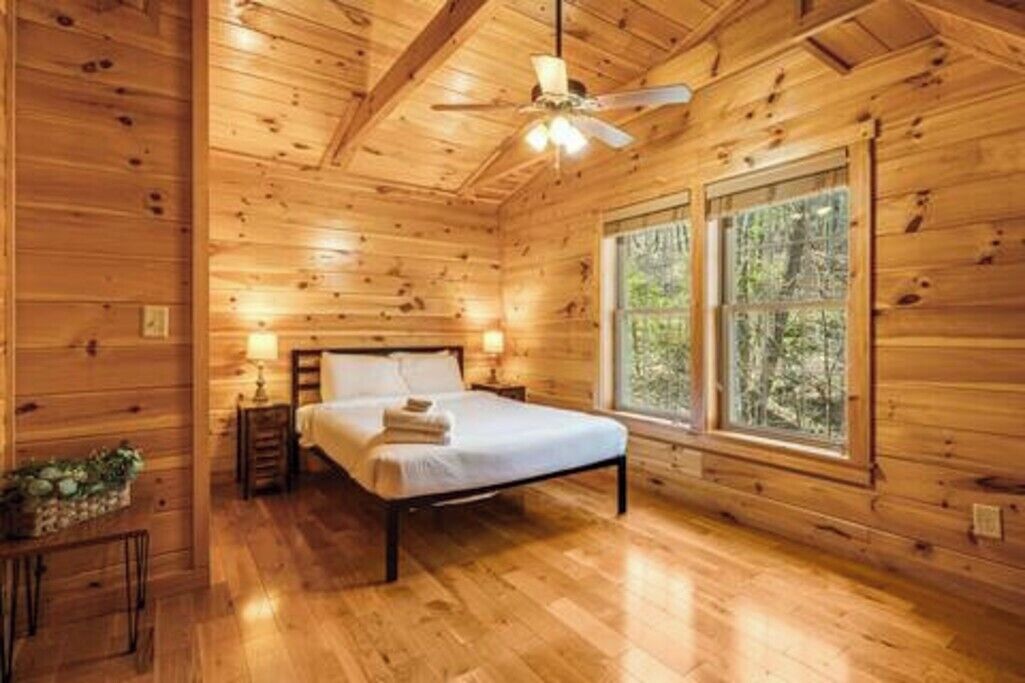 Une chambre à coucher dans une cabane en bois, avec un lit, un ventilateur de plafond et une fenêtre donnant sur la forêt.