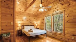 Een slaapkamer in een houten hutje, met een bed, plafondventilator en een raam met uitzicht op bomen.