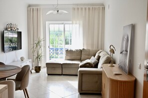A living room with a sofa, a wooden side table, a TV, and a window with curtains. - Sunny 2 bedroom close to beach (Albufeira)