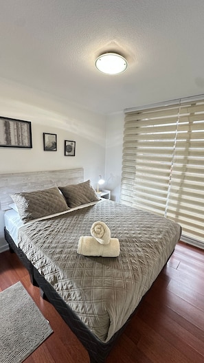 Een net opgemaakt bed met een gewatteerde spreidek, een opgerolde handdoek en een bedlampje.