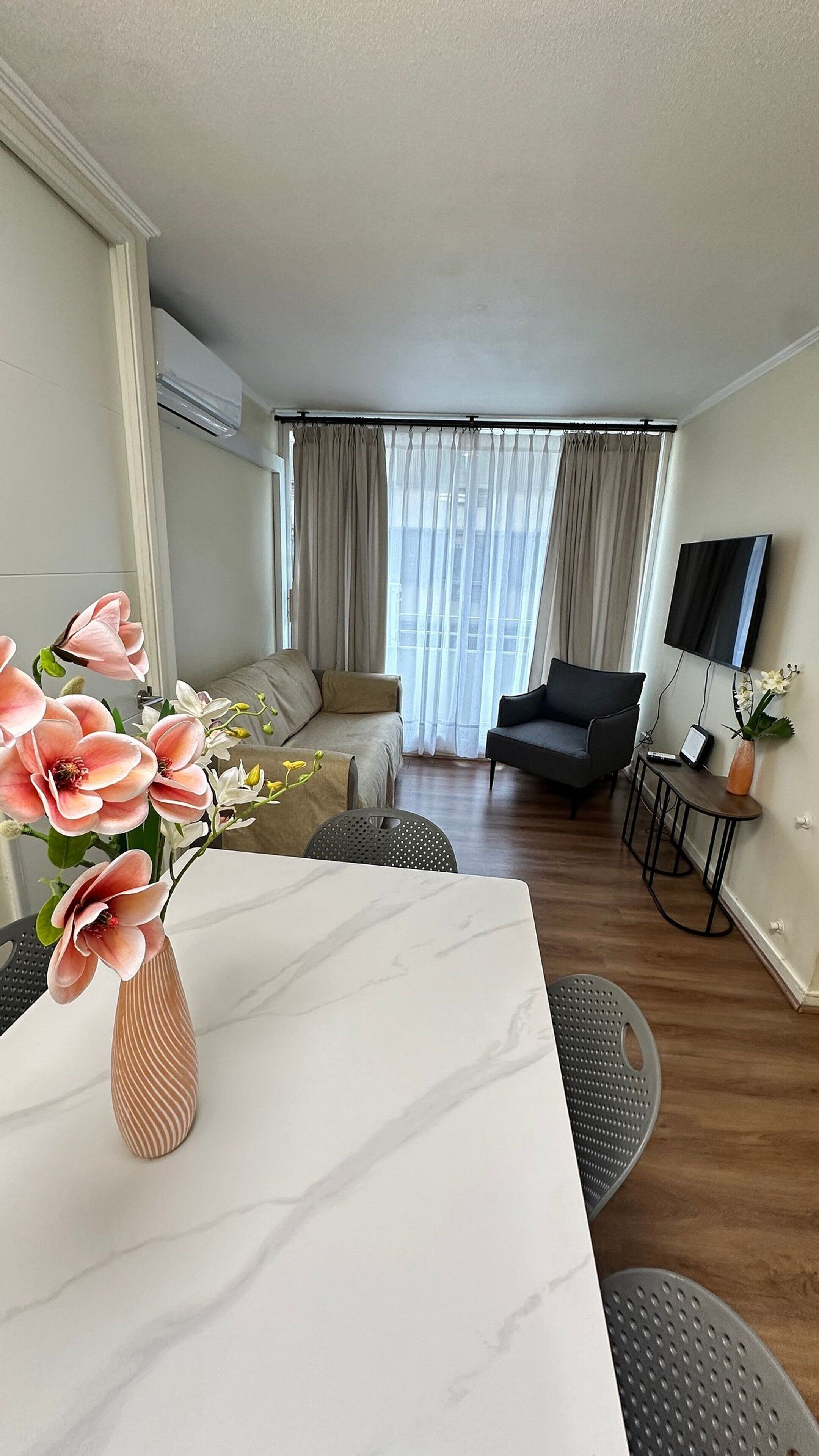 Ein modernes Wohnzimmer mit einer Couch, einem Esstisch mit Blumenvase, einem Fernseher und einem Fenster mit Vorhängen.