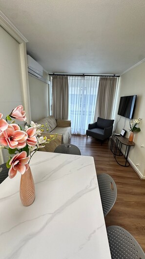 A modern living room with a sofa, a dining table with a vase of flowers, a TV, and a window with curtains. - Spacious apt. 2 bedr. Fully equipped downtown Stgo. (Santiago)